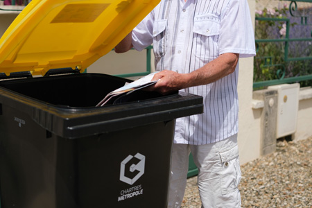 Une personne qui jette un papier dans un bac roulant jaune de Chartres métropole